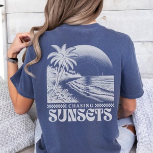 May include: A navy blue t-shirt with a white graphic of a palm tree on a beach with waves and the text "Chasing Sunsets" in a retro font.