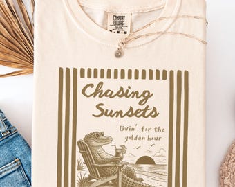 May include: A cream-colored t-shirt featuring a brown graphic print of an alligator wearing sunglasses, sitting in a beach chair with a drink. The text reads Chasing Sunsets and livin' for the golden hour. The shirt has a Comfort Colors brand label.