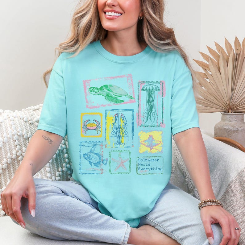 May include: A light blue t-shirt featuring a collection of sea creatures within colorful rectangular frames. The design includes a turtle, jellyfish, crab, lobster, fish, starfish, and a stingray. The text "Saltwater Heals Everything" is also printed on the shirt.
