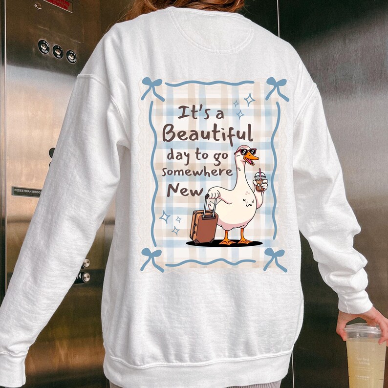 May include: White sweatshirt featuring a cartoon goose wearing sunglasses and holding a beverage, with a suitcase. The text reads "It's a Beautiful day to go somewhere New" in a blue and tan frame.