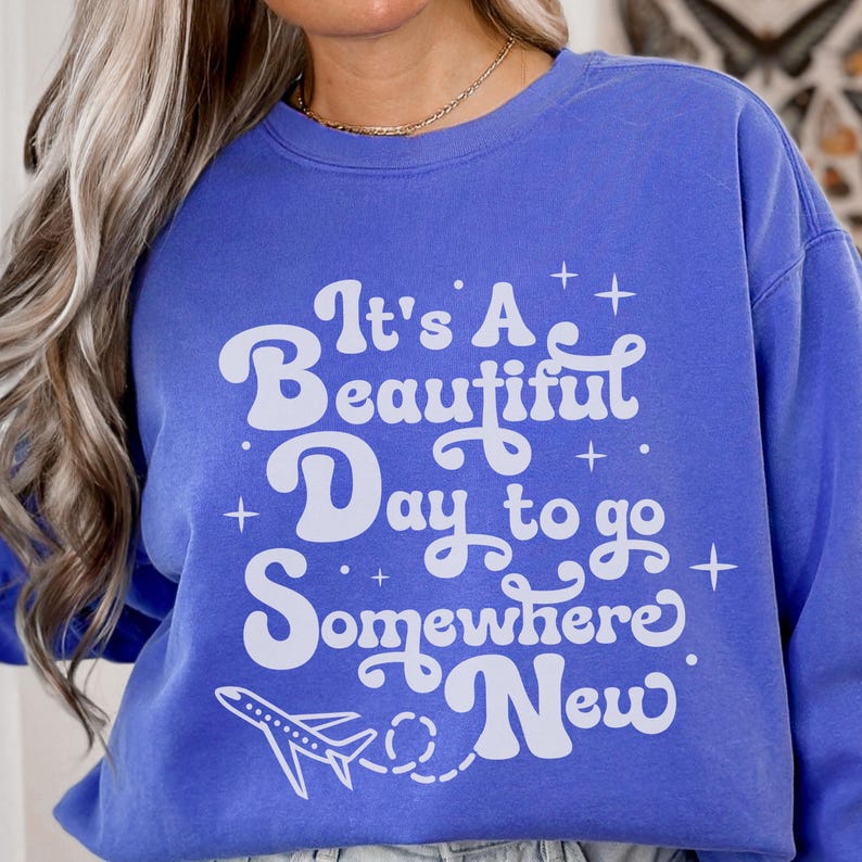 以下が含まれることがあります： 「It's A Beautiful Day to go Somewhere New」の白文字がプリントされた青色のスウェットシャツ。飛行機と点線でデザインされています。スウェットシャツは単色の青色です。