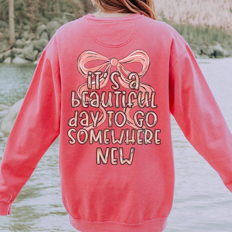 Puede incluir: Una sudadera de color coral con un gran gr&aacute;fico en la espalda. El gr&aacute;fico presenta un lazo y el texto "It's a beautiful day to go somewhere new" en una fuente l&uacute;dica. La sudadera tiene cuello redondo y mangas largas.