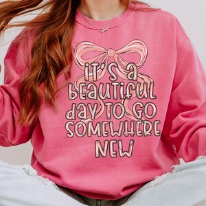Puede incluir: Sudadera rosa con la frase "It's a beautiful day to go somewhere new" en una fuente caprichosa, acentuada por un gr&aacute;fico de lazo. La sudadera es de color rosa liso.