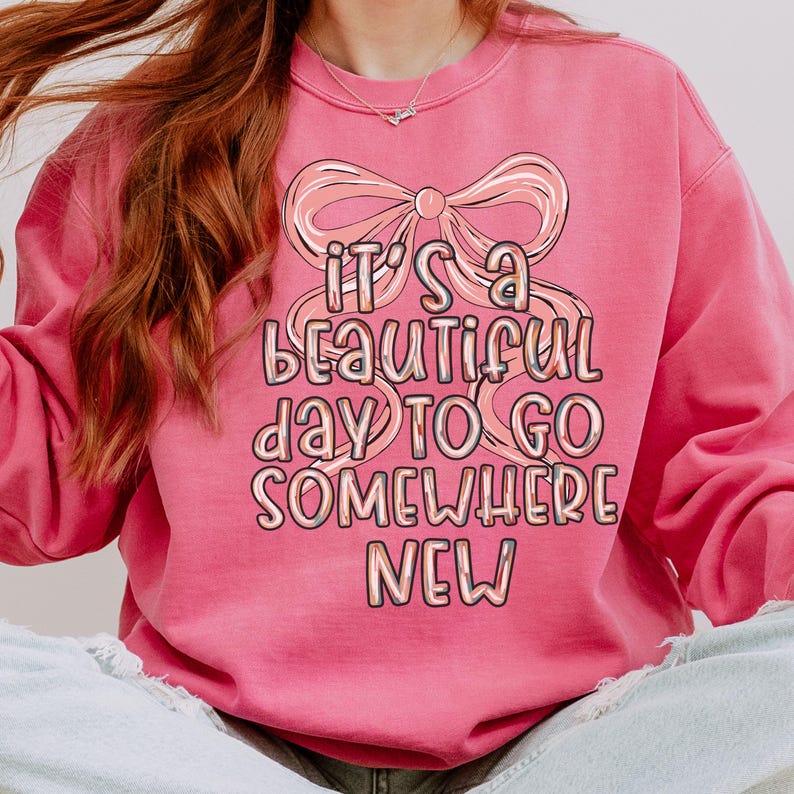 Puede incluir: Sudadera rosa con la frase "It's a beautiful day to go somewhere new" en una fuente caprichosa, acentuada por un gr&aacute;fico de lazo. La sudadera es de color rosa liso.
