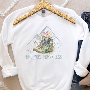 May include: White sweatshirt with a watercolor illustration of hiking boots on a mountain with flowers. The text "Hike More Worry Less" is printed below the illustration.