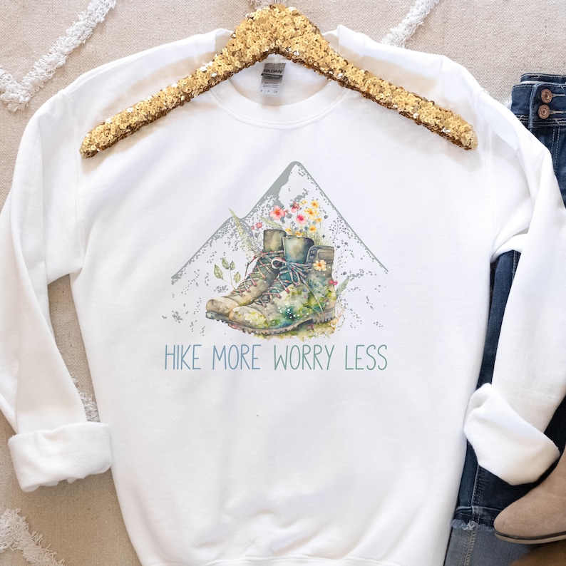 May include: White sweatshirt with a watercolor illustration of hiking boots on a mountain with flowers. The text "Hike More Worry Less" is printed below the illustration.