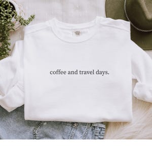 May include: White crew neck sweatshirt with the text "coffee and travel days." The sweatshirt is made of a soft material and is suitable for casual wear. The design is simple and minimalist.