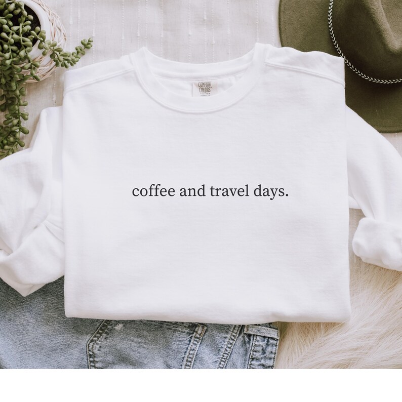 May include: White crew neck sweatshirt with the text "coffee and travel days." The sweatshirt is made of a soft material and is suitable for casual wear. The design is simple and minimalist.