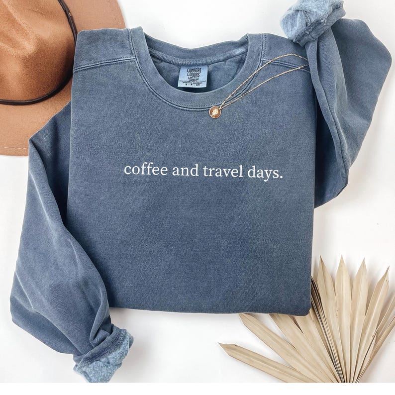 May include: A blue-grey sweatshirt with the text "coffee and travel days." A gold necklace with a pendant rests on the shirt. The sweatshirt has a crew neck and long sleeves with cuffed ends. A brown hat is visible in the background.