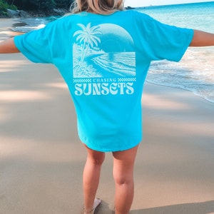 May include: A turquoise blue t-shirt with a white graphic of a palm tree and a beach scene. The text "Chasing Sunsets" is printed in white on the back of the shirt.