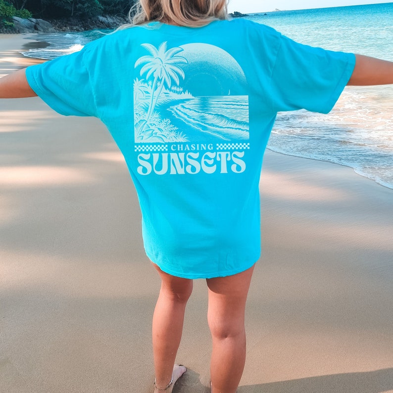May include: A turquoise blue t-shirt with a white graphic of a palm tree and a beach scene. The text "Chasing Sunsets" is printed in white on the back of the shirt.