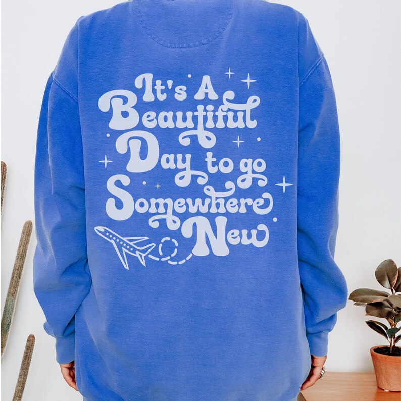 以下が含まれることがあります： 白い文字で「It's A Beautiful Day to go Somewhere New」と書かれた青いスウェットシャツ。デザインには小さな飛行機のグラフィックが含まれています。スウェットシャツは無地の青色です。