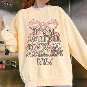 Puede incluir: Sudadera amarilla clara con un lazo rosa y el texto "It's a beautiful day to go somewhere new" en una fuente colorida. La sudadera tiene mangas largas y cuello redondo.