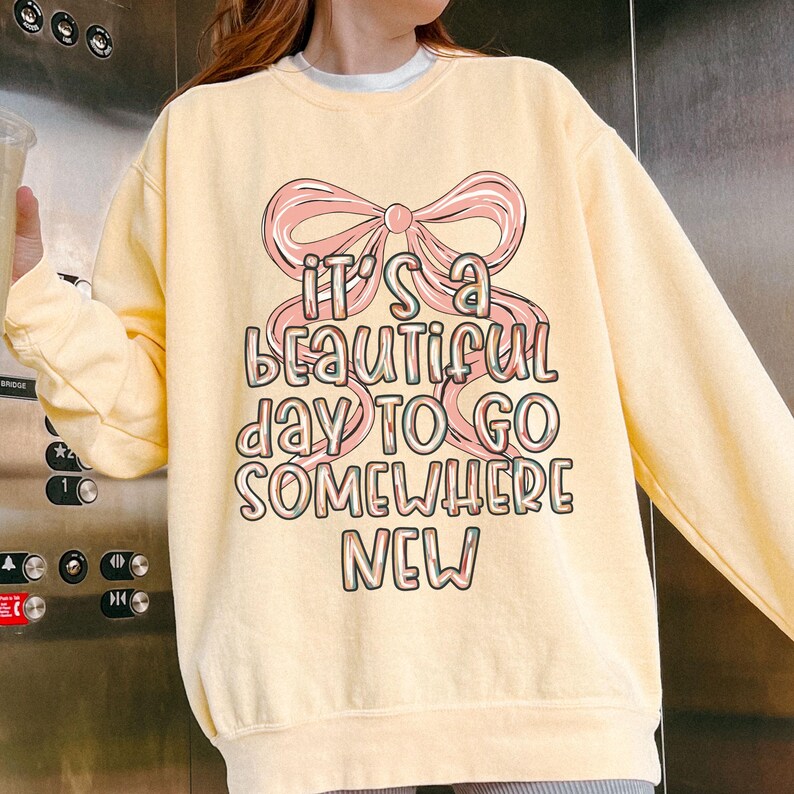 Puede incluir: Sudadera amarilla clara con un lazo rosa y el texto "It's a beautiful day to go somewhere new" en una fuente colorida. La sudadera tiene mangas largas y cuello redondo.