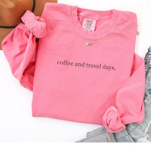 May include: A pink long-sleeved sweatshirt with the text "coffee and travel days." The shirt has a crew neck and is made of a soft material. A gold necklace with a small pendant is visible.