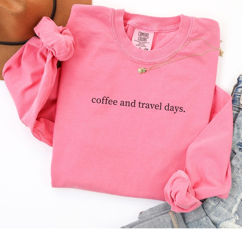 May include: A pink long-sleeved sweatshirt with the text "coffee and travel days." The shirt has a crew neck and is made of a soft material. A gold necklace with a small pendant is visible.