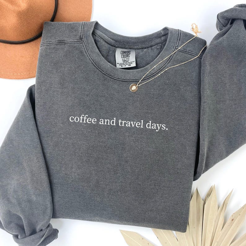 May include: A charcoal gray sweatshirt with the text "coffee and travel days" in white. A gold necklace with a small pendant is draped over the sweatshirt. A brown hat is visible in the background.
