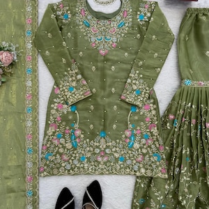 May include: An olive green embroidered salwar kameez set with matching dupatta and bottoms. The set features intricate floral embroidery in pink, blue, and gold. Black slip-on shoes with silver accents are at the bottom. A floral necklace is also present.
