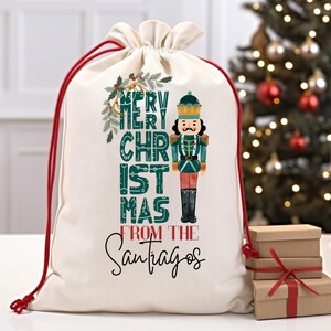 May include: A white canvas gift bag with red drawstrings. The bag features a green and white illustration of a nutcracker and the text "Merry Christmas from the Santiagos".