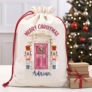 May include: A white canvas Christmas gift bag with red drawstring. The bag features a watercolor illustration of a red door with a wreath, two nutcracker figurines, and the text "Merry Christmas Adrian".