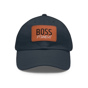 Pode incluir: Boné de beisebol azul marinho com um patch de couro castanho. O patch apresenta as palavras "BOSS Mindset" em escrita branca. O boné tem um design clássico de seis painéis.