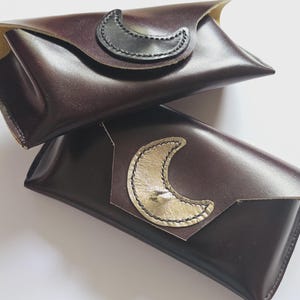 May include: Two dark brown leather eyeglass cases. Each case has a flap closure with a crescent moon accent. One moon is gold, the other is black. The cases are stacked on top of each other.