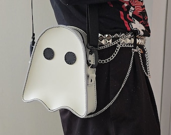 May include: A white ghost-shaped crossbody bag with black eyes. The bag is worn over a black shirt with a silver studded belt and a silver chain.