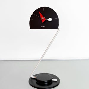 May include: A modern black quartz clock with a red pointer and white hands. The clock face is semi-circular and mounted on a white metal stand. The base is a black circular disc. The word "QUARTZ" is printed on the clock face.