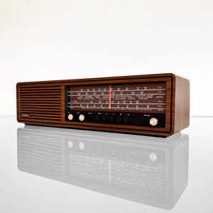 Könnte beinhalten: Ein rechteckiges SABA-Radio mit dunkler Holzmaserung, das links ein horizontales Lautsprechergitter und rechts ein schwarzes Glasdisplay aufweist. Das Display zeigt Frequenzskalen, weiße Schrift und einen vertikalen roten Abstimmungsanzeiger.