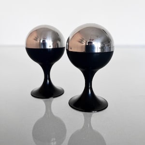 May include: A pair of spherical salt and pepper shakers featuring a polished silver-tone metal top half and a matte black base. Each shaker is supported by a slender, flared pedestal stand, creating a balanced, rounded silhouette for table use.