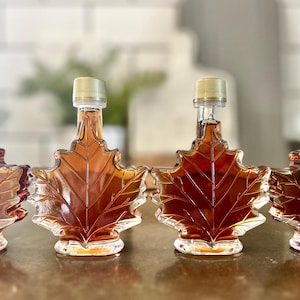 May include: Four clear glass bottles shaped like maple leaves filled with amber-colored maple syrup. The bottles have silver lids.