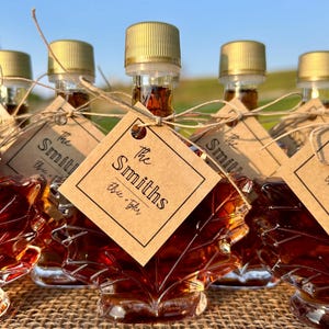 May include: Several maple syrup bottles shaped like maple leaves. Each bottle has a gold cap and a brown tag that reads "The Smiths" with "Elsie + Tyler" below. The bottles are tied with twine and sit on a burlap surface.