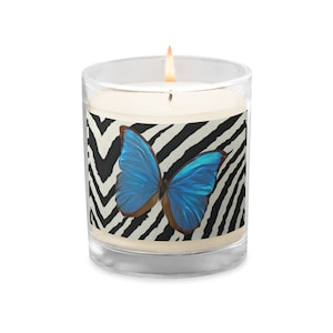 Puede incluir: Un tarro de vidrio para velas con una vela blanca y un gráfico de mariposa azul sobre un fondo de estampado de cebra blanco y negro.