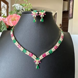 May include: A gold-tone necklace and matching stud earring set featuring marquise-cut stones in vibrant green, bright pink, and white. The necklace has a V-shaped design with a central floral motif and a dangling green teardrop stone.
