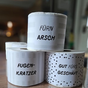 May include: White rolls of paper with black text. One roll says "F&Uuml;RN ARSCH", another says "FUGEN-KRATZER", and the third says "GUT F&Uuml;RS GESCH&Auml;FT". The rolls have different patterns, including stripes and polka dots.