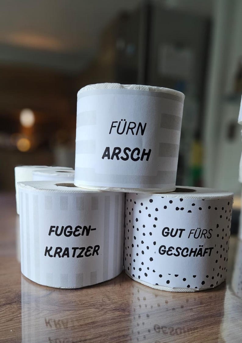 May include: White rolls of paper with black text. One roll says "F&Uuml;RN ARSCH", another says "FUGEN-KRATZER", and the third says "GUT F&Uuml;RS GESCH&Auml;FT". The rolls have different patterns, including stripes and polka dots.