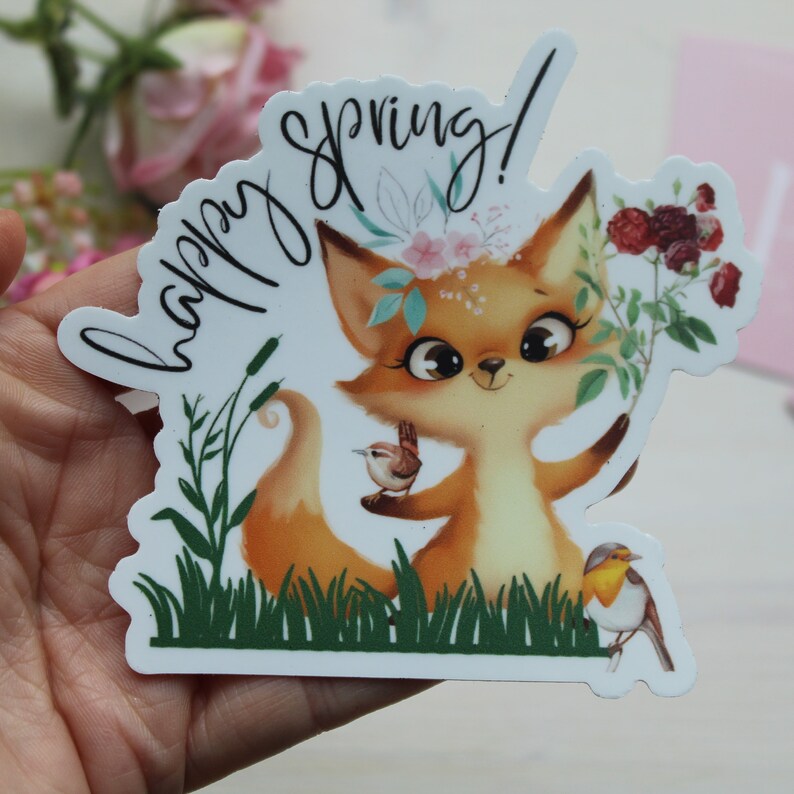 May include: A sticker featuring a cartoon fox with a floral crown, holding flowers and a bird. The sticker has the words "happy spring!" in a cursive font. The fox is orange and white, with large eyes and a smiling expression. The sticker is surrounded by green grass and other birds.