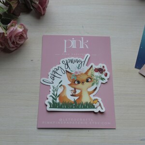 May include: A sticker featuring a cartoon fox with a floral crown, holding flowers and a bird, with the words "happy spring!" in a cursive font. The sticker is on a pink card with the text "pink fine papeterie".
