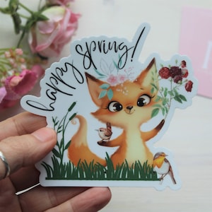 May include: A sticker featuring a cartoon fox with a floral crown, holding flowers and a bird. The sticker has the words "happy spring!" in a script font. The fox is orange and white, with big eyes and a smiling expression.