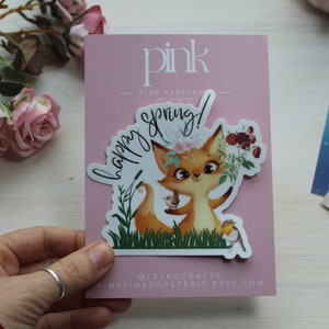May include: A sticker featuring a cartoon fox with a floral crown, holding a teacup, and surrounded by flowers and a bird. The sticker is on a pink card with the words "happy spring!" and "pink fine papeterie".