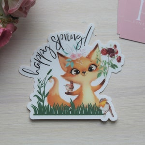 May include: A sticker featuring a cartoon fox with a floral crown, holding a bird. The words "happy spring!" are written in cursive. The fox is surrounded by green grass and flowers, with a white background.