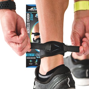 May include: Black Achilles tendon strap with a triangular design, being adjusted on an ankle. The strap is labeled "ACHILLES TENDON STRAP" and "SMALL: 10"-14"". The person is wearing a black watch and a yellow wristband. The strap is being adjusted over a black and gray athletic shoe.
