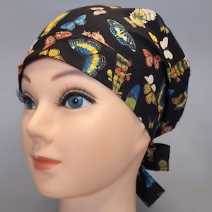 Puede incluir: Un gorro de quirófano negro con estampado de mariposas de colores. El gorro tiene un lazo en la parte posterior.