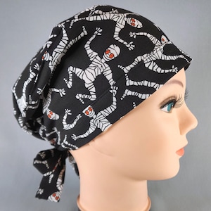 Puede incluir: Gorro quirúrgico negro con estampado de momias blancas y ojos rojos. El gorro está atado en la parte posterior y se muestra en una cabeza de maniquí. La tela es de color negro oscuro con un patrón repetitivo de momias.