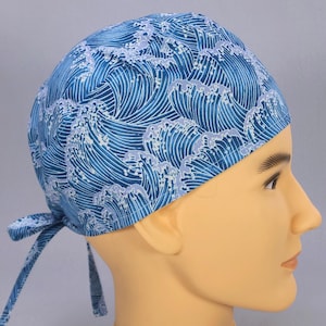 May include: A blue surgical scrub cap with a repeating wave pattern in shades of blue and white. The cap is tied at the back with two fabric ties. The cap is displayed on a mannequin head.