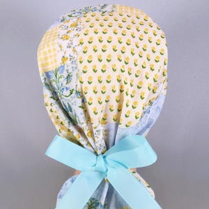 May include: A patterned scrub cap with a light blue bow. The cap features a patchwork design with yellow, blue, and white floral and gingham patterns. The bow is tied at the base of the cap.