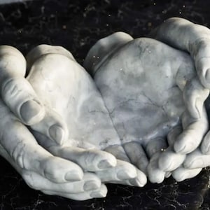 Może przedstawiać: Rzeźba z jasnoszarego kamienia przedstawiająca dwie dłonie złożone w kształt misy. Dłonie mają widoczne żyły i pęknięcia, co nadaje im wygląd postarzenia. Rzeźba spoczywa na ciemnej, marmurowej powierzchni.