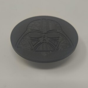 May include: A dark gray, circular object featuring a detailed relief of Darth Vader's helmet. The design is centered on the disc, showcasing the character's iconic mask. The object appears to be made of a solid material.