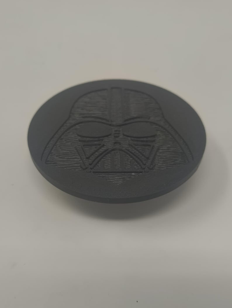 May include: A dark gray, circular object featuring a detailed relief of Darth Vader's helmet. The design is centered on the disc, showcasing the character's iconic mask. The object appears to be made of a solid material.