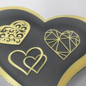 May include: A heart-shaped decorative tray with a black interior and gold trim. The tray features three gold heart designs: a filigree heart, a geometric heart, and two intertwined hearts. The tray is likely used for jewelry or small items.